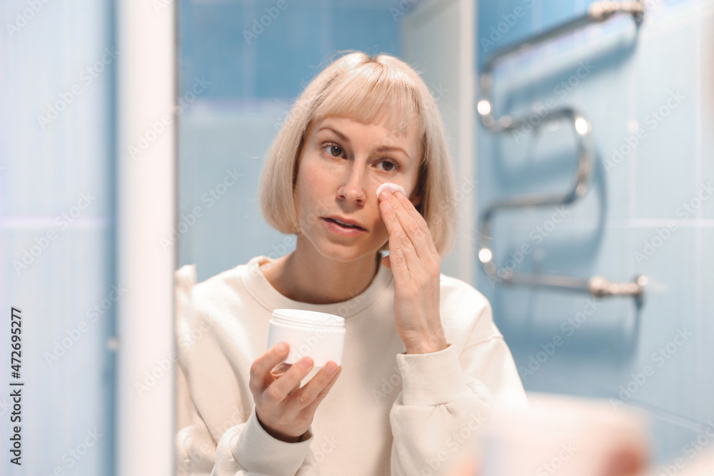 Beautiful young woman applying moisturizer on her face in bathroom, taking care of her skin Beautiful young woman applying moisturizer on her face in bathroom, taking care of her skin