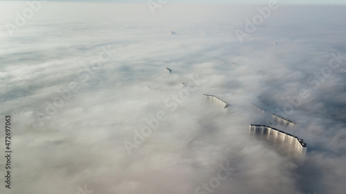 Fototapeta Naklejka Na Ścianę i Meble -  Silhouettes of tall houses through the incoming thick fog. Megalopolis in white clouds in the early morning at time.