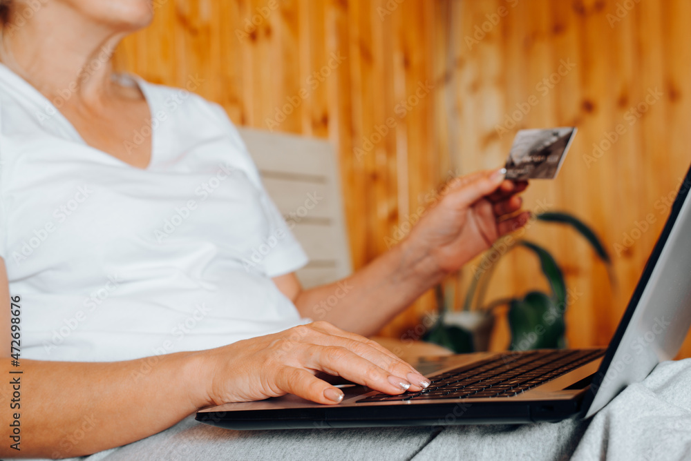 Naklejka premium Side view of woman typing on laptop and holding bank credit or debit card while sitting on bed indoors, close-up. Online shopping, e-commerce concept. Selective focus in the foreground