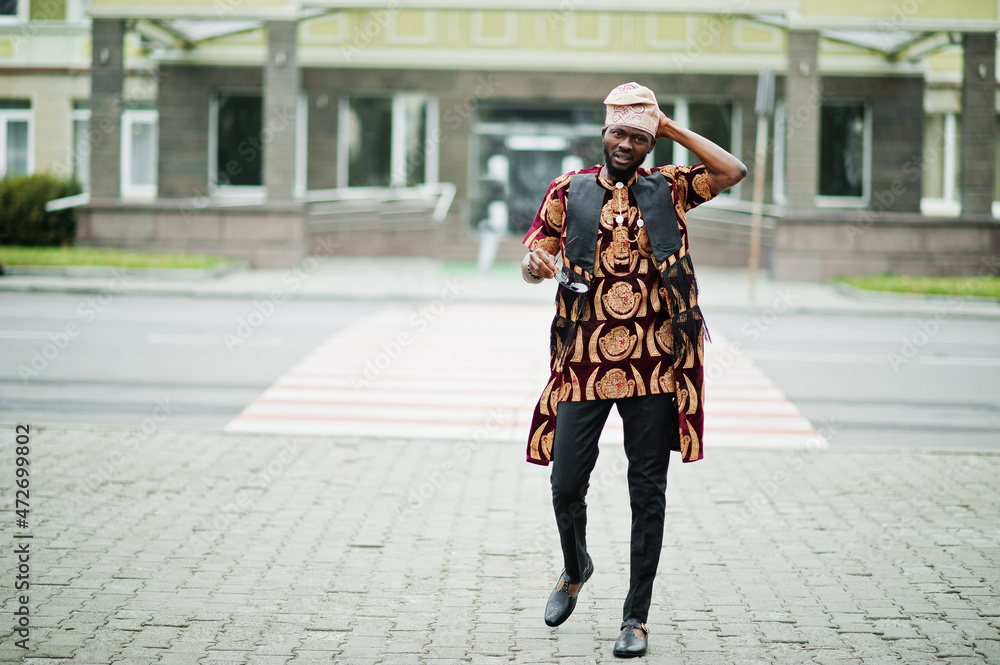 African stylish and handsome man in traditional outfit and cap standing outdoor.