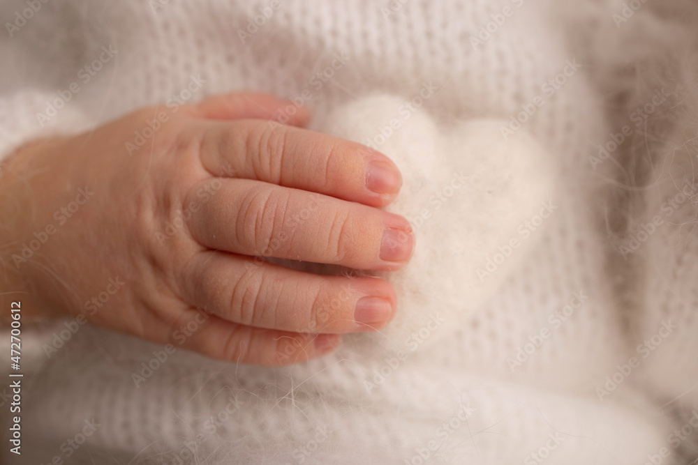 Tiny hand of a newborn baby. Soft hands of new born in white clothes hold white woolen heart. Fingers of a newborn baby hands close up. Studio macro photo. Happiness of a woman. Photography, concept. Tiny hand of a newborn baby. Soft hands of new born in white clothes hold white woolen heart. Fingers of a newborn baby hands close up. Studio macro photo. Happiness of a woman. Photography, concept.