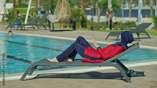 Stylish young woman wearing hijab swimsuit and sunglasses sunbathing by the pool.Happy woman enjoying vacation and sun by the pool.
