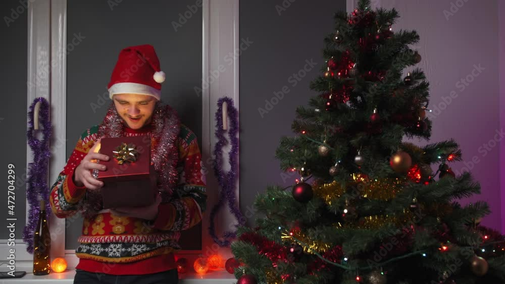 A young man opens a gift from which a light shines, he is wearing a Christmas sweater and a hat, next to him is a New Year tree, a concept of Christmas and New Year A young man opens a gift from which a light shines, he is wearing a Christmas sweater and a hat, next to him is a New Year tree, a concept of Christmas and New Year