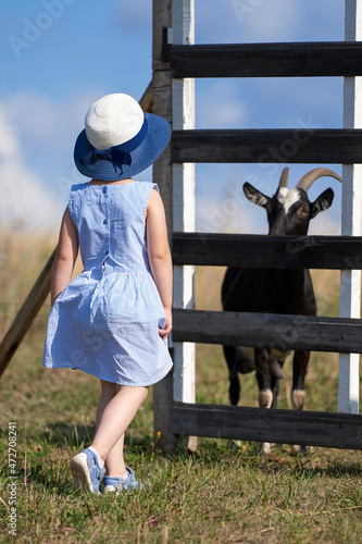 A little girl is standing at the corral with goats. The girl and the goat look at each other.