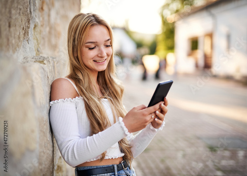 Cheerful teenager student girl standing in front of a vintage stone wall and orering something online with a smart phone - Young woman checking her social media feed and sharing a story