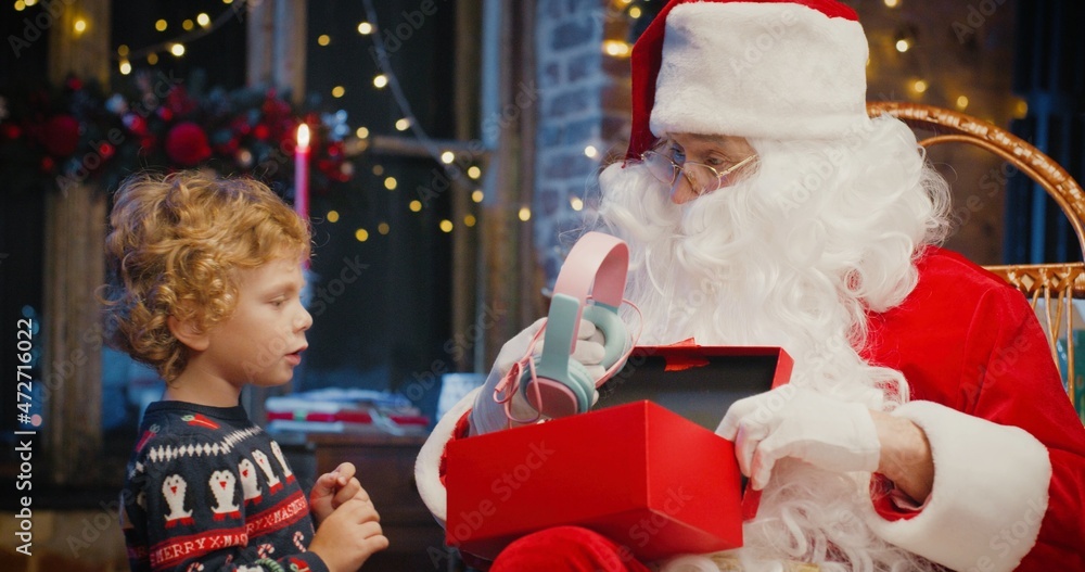 Santa Claus and little caucasian boy standing nearby and waiting for magical Christmas gift near the Christmas tree in cozy living room in winter Santa Claus and little caucasian boy standing nearby and waiting for magical Christmas gift near the Christmas tree in cozy living room in winter