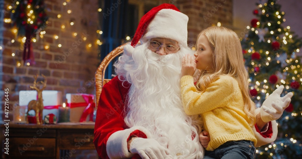 I will tell you but keep it in secret. Portrait view of the little blonde girl sitting at the Santa knees while sharing secrets with him I will tell you but keep it in secret. Portrait view of the little blonde girl sitting at the Santa knees while sharing secrets with him
