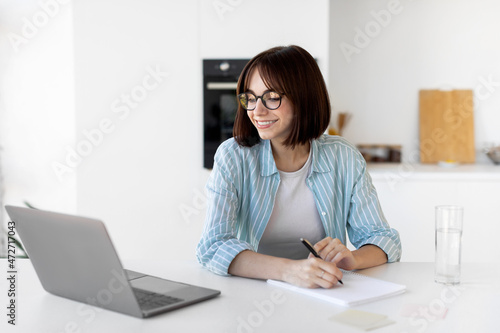 Young freelancer woman working with laptop in kitchen and writing down information while sitting in kitchen