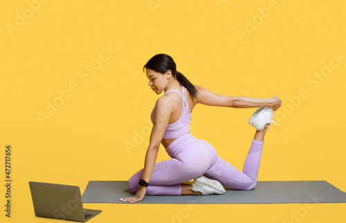 Fitness online. Young black woman stretching, sitting on yoga mat in front of laptop, exercising over yellow background