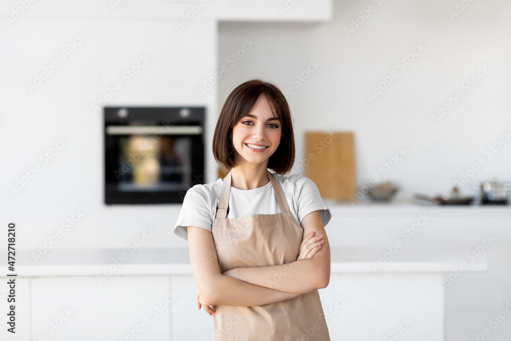 Young caucasian lady in apron standing with folded arms, looking and smiling at camera, posing in kitchen Young caucasian lady in apron standing with folded arms, looking and smiling at camera, posing in kitchen