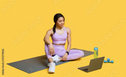 Ready for training. Fit african american lady browsing fitness tutorial on laptop, sitting on mat, yellow background
