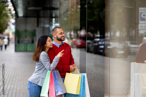 Portrait of smiling young inrerracial couple holding shopper bags, pointing at shop window, walking near shopping mall