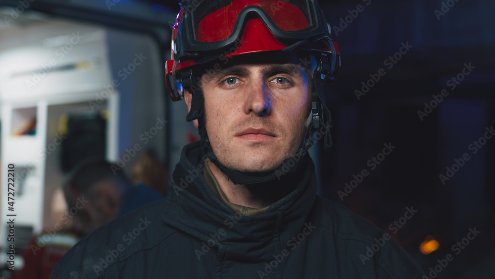 Man in hardhat looking at camera while standing near ambulance van with paramedics and survivor after emergency at night Man in hardhat looking at camera while standing near ambulance van with paramedics and survivor after emergency at night