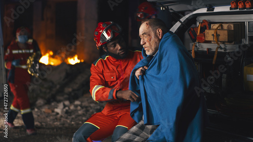 African American bearded man in paramedic uniform asking questions to senior man wrapped in blanket after accident at night photo