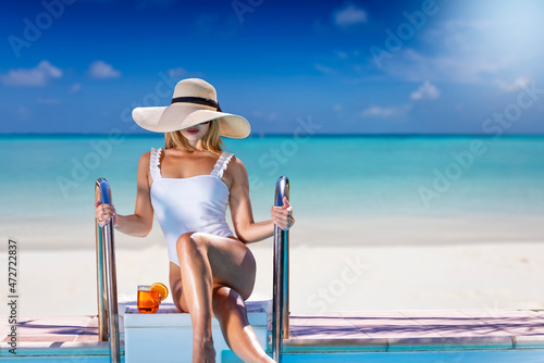 A attractive, blonde woman in a swimsuit and with sunhat sits by the swimming pool with a drink in front of a tropical paradise beach with turquoise sea