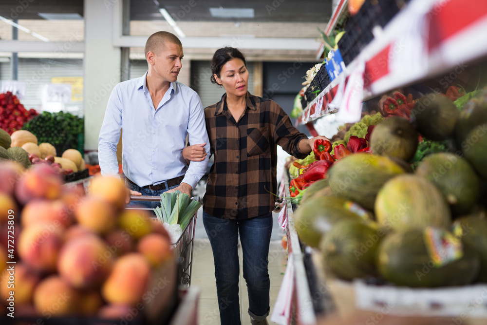 Obraz premium Pair of European man and Asian woman choosing vegetables and fruits in greengrocer.
