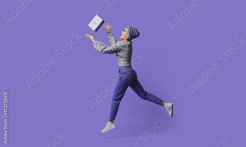 Full length side view photo of shocked caucasian woman wearing knitted hat and sweater catching gift box. Isolated over purple background