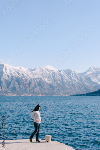 Pregnant woman stands on a pier against the backdrop of mountains and sea