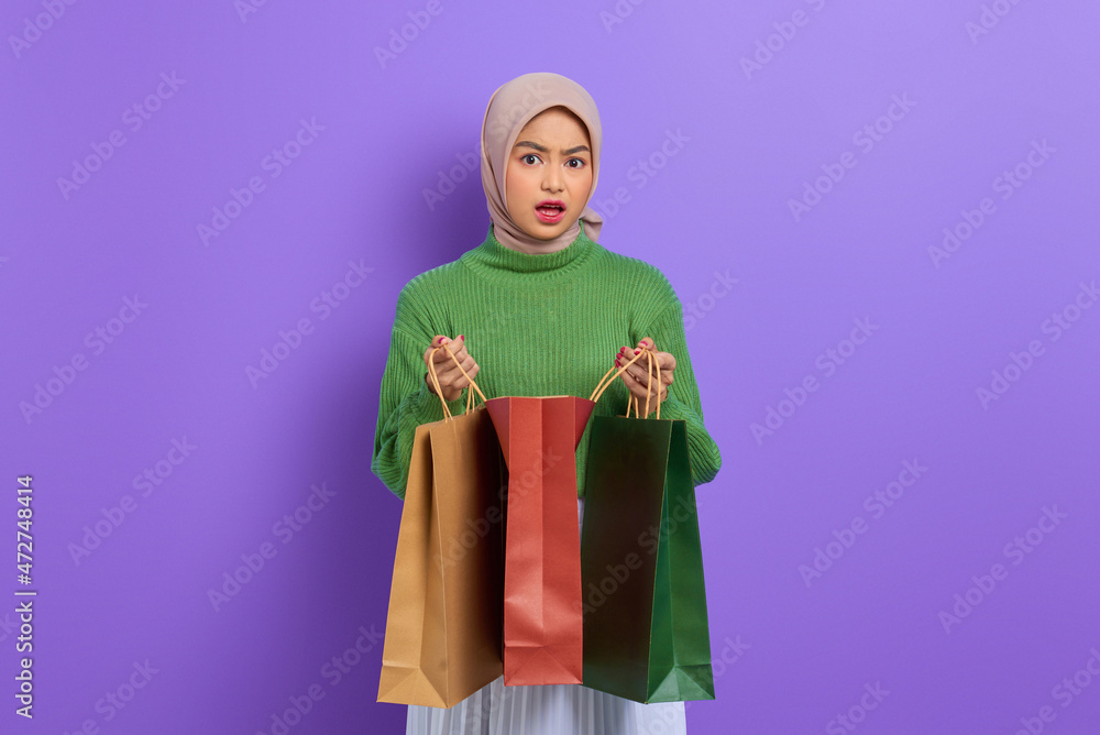 Shocked beautiful Asian woman in green sweater opening shopping bags isolated over purple background
