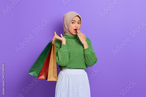 Shocked beautiful Asian woman in green sweater holding shopping bags with hand on mouth isolated over purple background