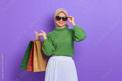Cheerful beautiful Asian woman in green sweater and sunglasses holding shopping bags isolated over purple background