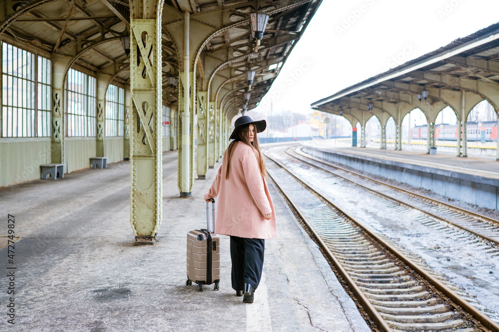 Young woman in coat walks with suitcase along empty platform of railway station. Caucasian girl tourist. Alone travel concept