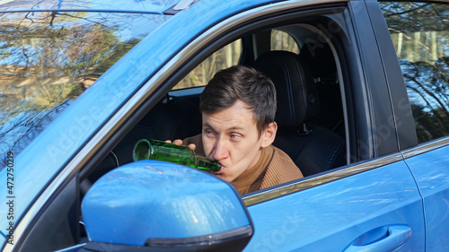 Driver brunet short-haired man sits in car cabin and drinks alcohol from glass bottle secretly hiding from police after hard work day, parked by forest photo
