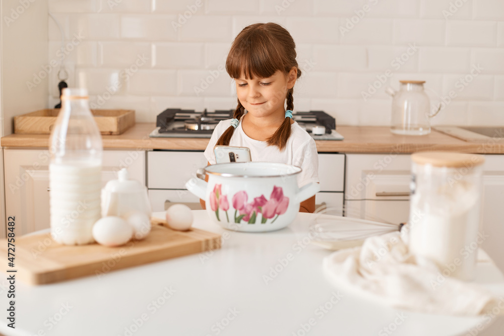 Portrait of charming cute dark haired female kid with pigtails sitting at table and using smart phone, finding recipe in the internet how to bake cake or cookies. Portrait of charming cute dark haired female kid with pigtails sitting at table and using smart phone, finding recipe in the internet how to bake cake or cookies.
