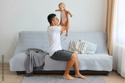 Indoor shot of ghandsome brunette father holding infant baby girl in hands and understand they need changing baby's diaper, feeling bad smell, raising kid up air.