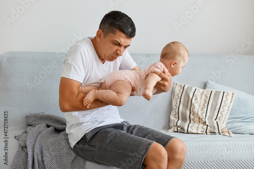 Portrait of frustrated man posing with toddler girl in hands, smelling baby, needs to change diaper and wash kid, sitting on sofa in light living room at home.