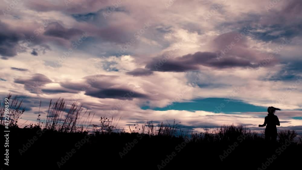 Beautiful black silhouette outline of a female jogger, jogging against a dramatic sky.  The jogger runs toward the camera from right to left.  A wide angle frames this stunning clouds and scenery.