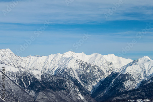 Snowy mountain ranges on the background of the blue sky with clouds. The lower part of the mountains is black with only a little bit of snow, while the tops are completely white.