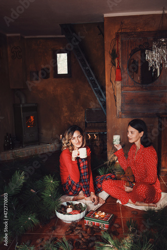 Two young women in red pajamas drinking coffee in cottage, december morning, christmass decoration