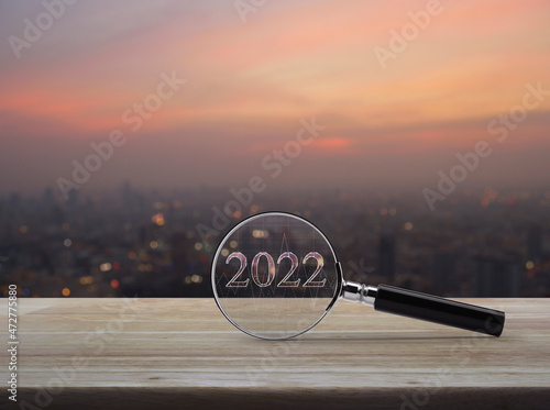 Magnifying glass with 2022 text and financial graph on wooden table over blur of cityscape on warm light sundown, Business happy new year 2022 research concept
