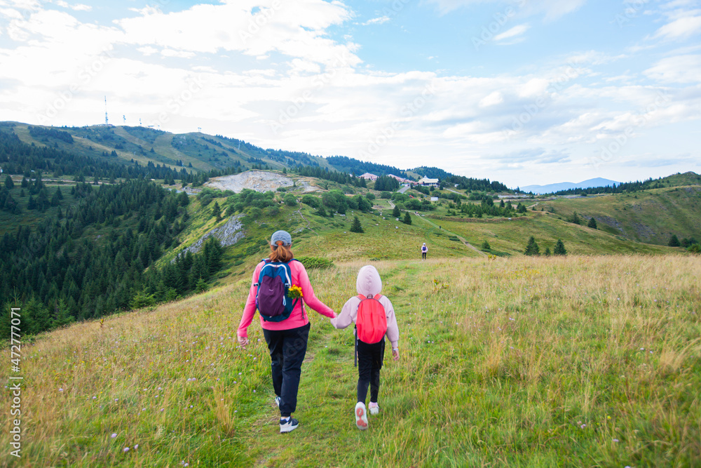 Mother and daughter with backpack hike on mountain trail, beautiful summer day, mountain nature landscape. Family outdoor activity.