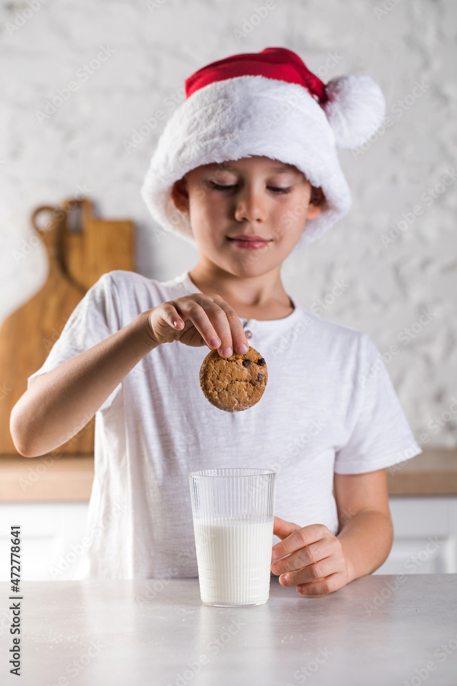 Cute little boy in Santa hat and white t-shirt puts cookie into milk in kitchen
