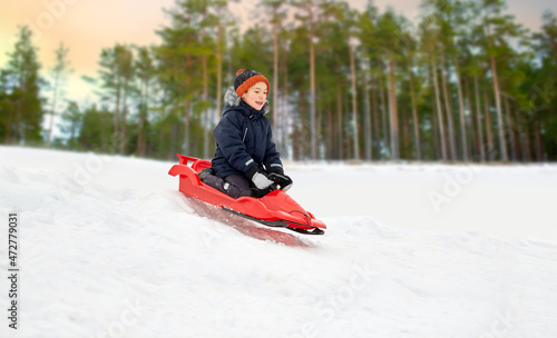 childhood, sledging and season concept - happy little boy sliding on sled down snow hill outdoors in winter over snowy forest or park background
