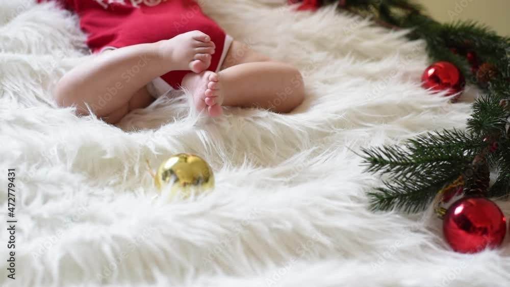 Legs of baby in red jumpsuit lying on white blanket next to Christmas tree branches and balls. Kid moves foots. Close-up. Christmas and New year - beautiful and wonderful holidays. Legs of baby in red jumpsuit lying on white blanket next to Christmas tree branches and balls. Kid moves foots. Close-up. Christmas and New year - beautiful and wonderful holidays.