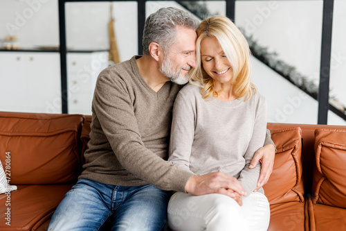Billede på lærred Gentle and lovely middle-aged couple in love sitting in embrace on the comfortable sofa at home, looks to each other tenderly