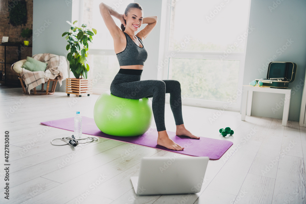 Full length body size view of attractive cheerful slim thin girl doing yoga stretching sitting on ball watching video at home indoors Full length body size view of attractive cheerful slim thin girl doing yoga stretching sitting on ball watching video at home indoors
