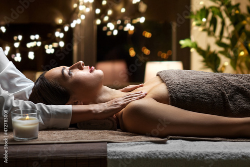 wellness, beauty and relaxation concept - beautiful young woman having face and head massage at spa over christmas lights on window background