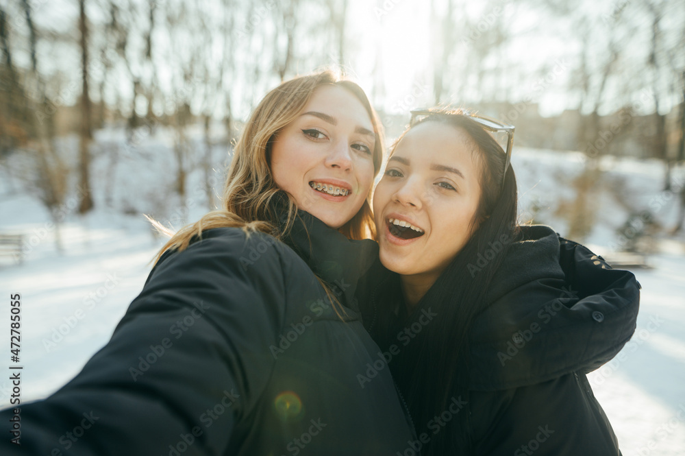 Two happy girlfriends in warm clothes take a selfie while walking in a snowy forest in sunny weather, look into the camera and smile. Two happy girlfriends in warm clothes take a selfie while walking in a snowy forest in sunny weather, look into the camera and smile.