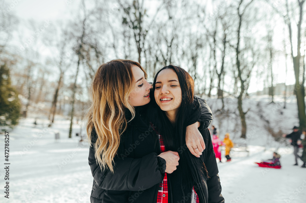 Two happy beautiful girlfriends hugging and smiling in a snowy forest on a walk. Leisure of friends in the winter season on a walk. Two happy beautiful girlfriends hugging and smiling in a snowy forest on a walk. Leisure of friends in the winter season on a walk.