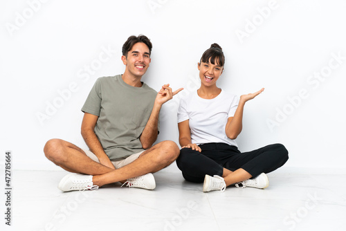 Young mixed race couple sitting on the floor isolated on white background pointing finger to the side and presenting a product
