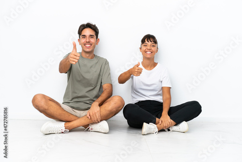 Young mixed race couple sitting on the floor isolated on white background giving a thumbs up gesture because something good has happened