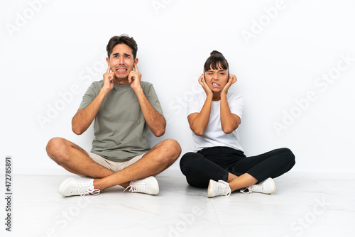 Young mixed race couple sitting on the floor isolated on white background covering both ears with hands