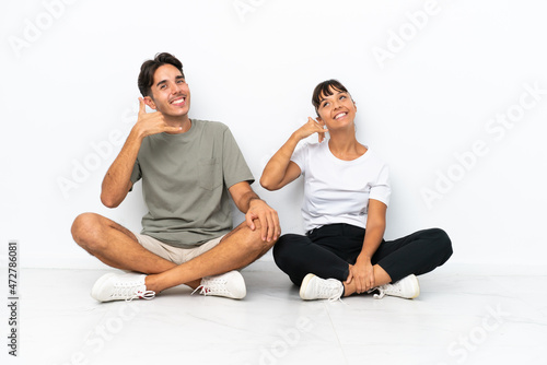Young mixed race couple sitting on the floor isolated on white background making phone gesture. Call me back sign