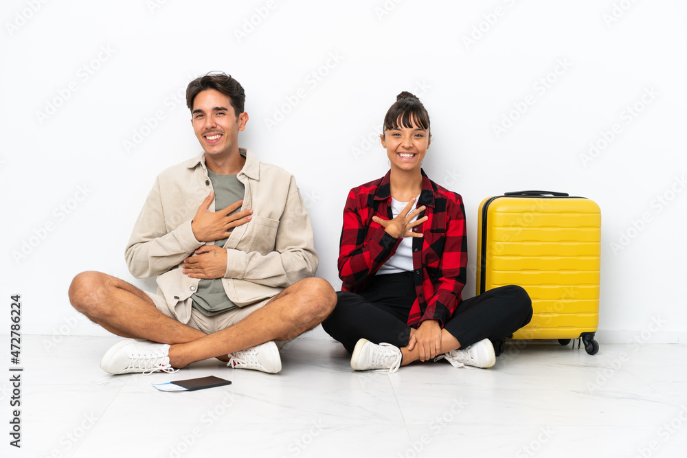 Young mixed race travelers couple sitting on the floor isolated on white background smiling a lot while putting hands on chest Young mixed race travelers couple sitting on the floor isolated on white background smiling a lot while putting hands on chest