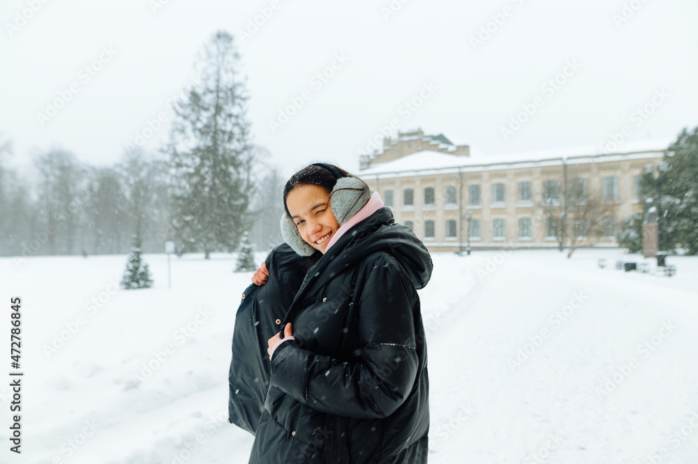 Positive hispanic lady in a black coat stands in a winter park and poses for the camera with a smile on her face in the snowfall. Positive hispanic lady in a black coat stands in a winter park and poses for the camera with a smile on her face in the snowfall.