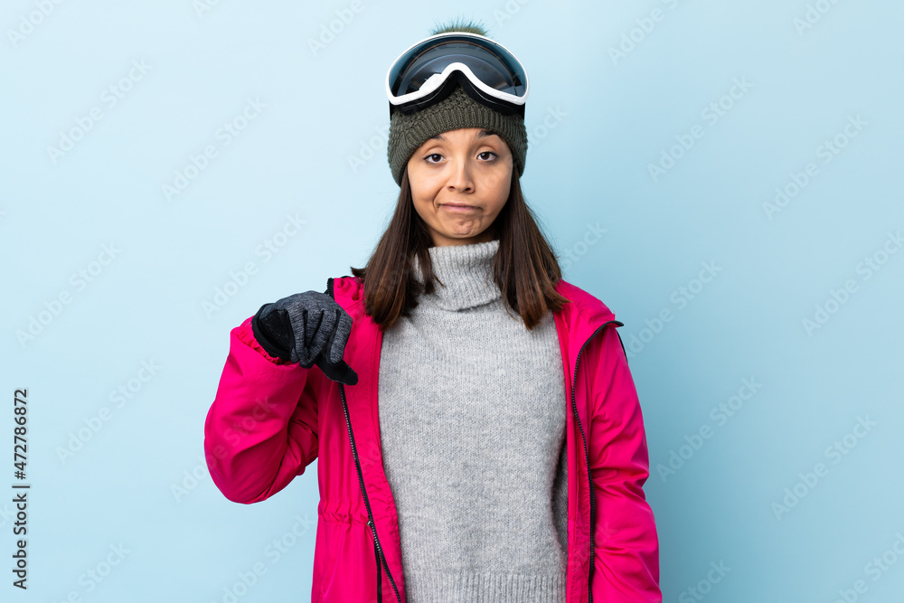 Mixed race skier girl with snowboarding glasses over isolated blue background showing thumb down with negative expression. Mixed race skier girl with snowboarding glasses over isolated blue background showing thumb down with negative expression.